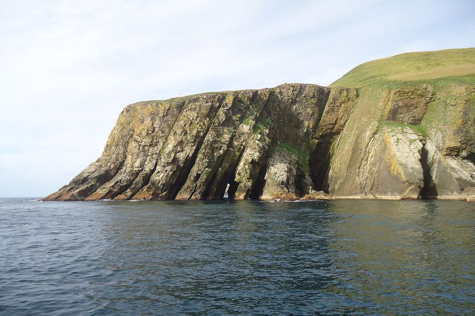 Tilted layers of ochre and grey sandstone form a cliff with dark sea arches at its base, a green grassy slope rising at right, over calm grey water