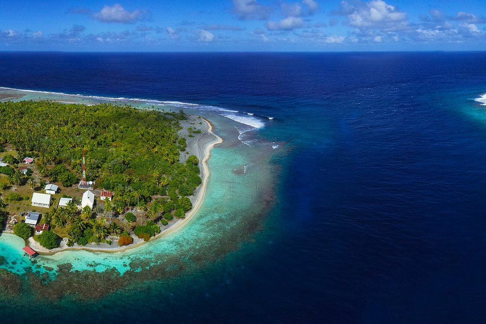 An aerial view of a palm-covered islet with a small village and jetties beside a deep blue channel through a turquoise reef