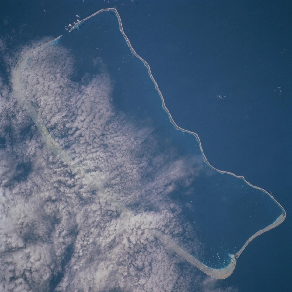 A photograph from orbit of a long rectangular ring of reef enclosing a dark blue lagoon, half covered by white cloud