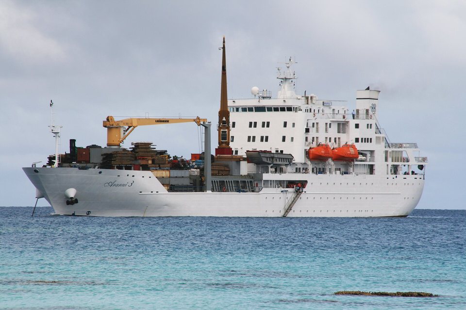 A white cargo-passenger ship with deck cranes and stacked cargo anchored on turquoise water under grey cloud