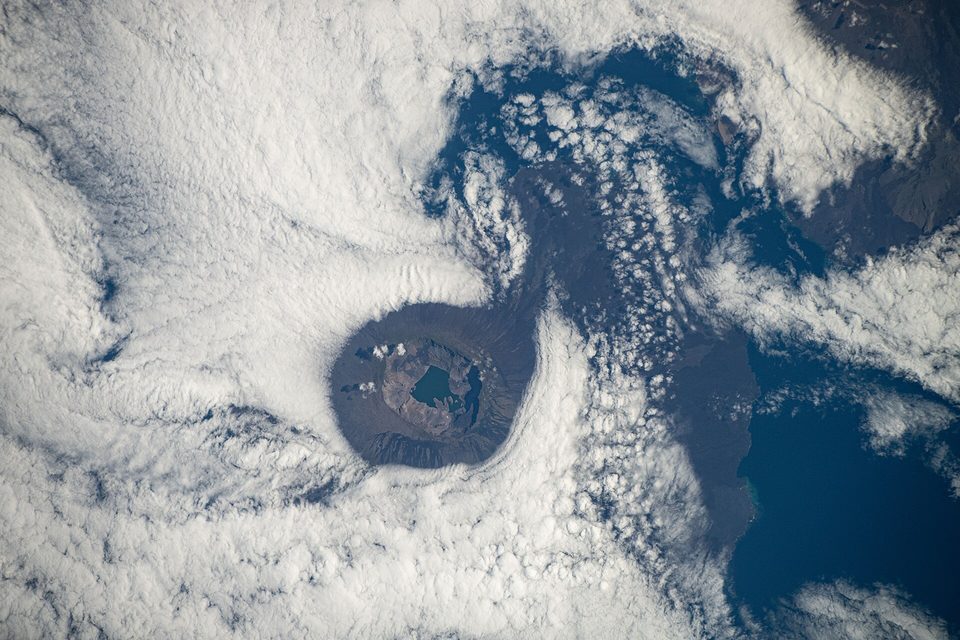 A round volcano with a wide caldera seen from orbit, clouds swirling around its flanks and the dark ocean beyond