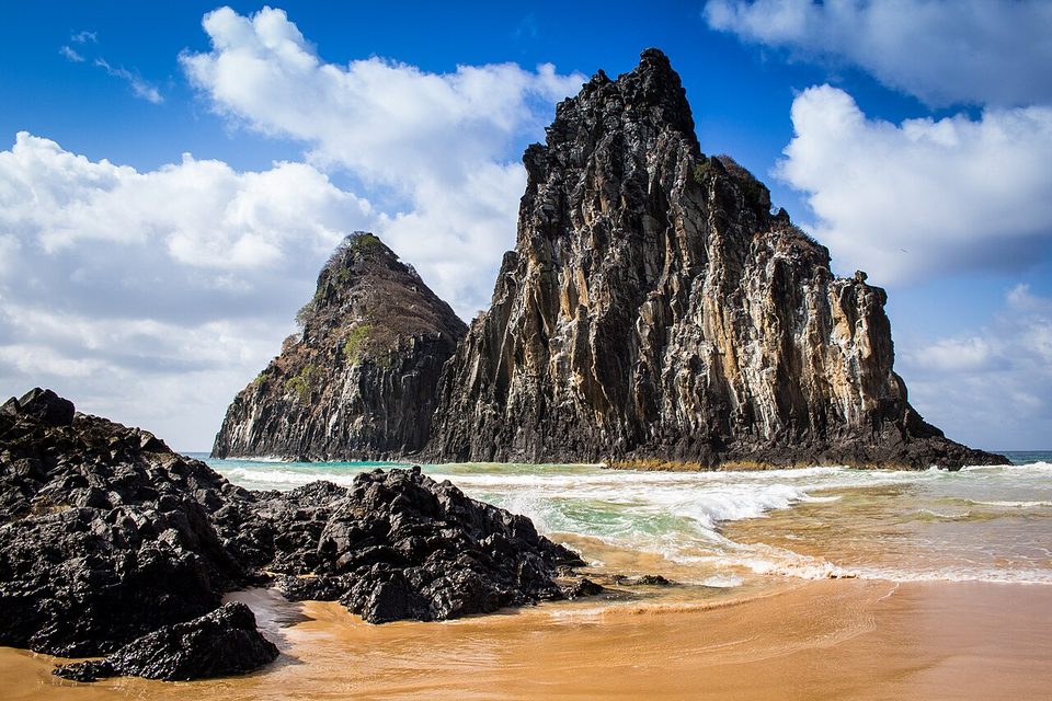 Two steep rock peaks rise from surf behind dark boulders on a golden beach under white clouds