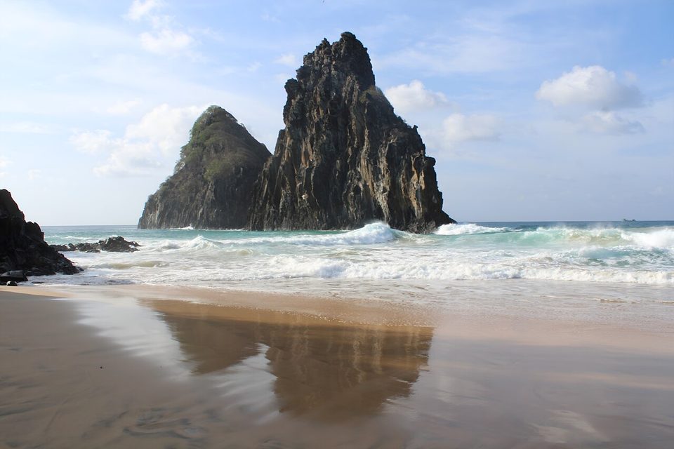 The twin peaks stand offshore in breaking surf, their reflection on wet sand in the foreground