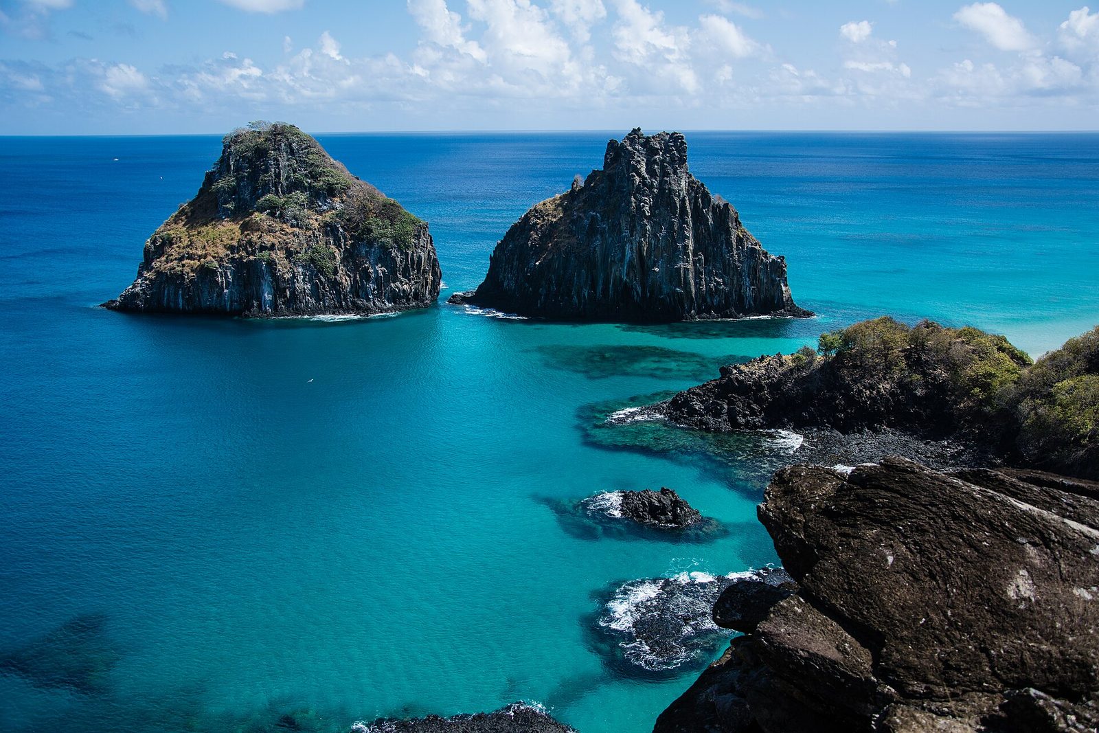 Two dark rock peaks stand in vivid turquoise water seen from the overlook, with reef rocks at right and ocean to the horizon