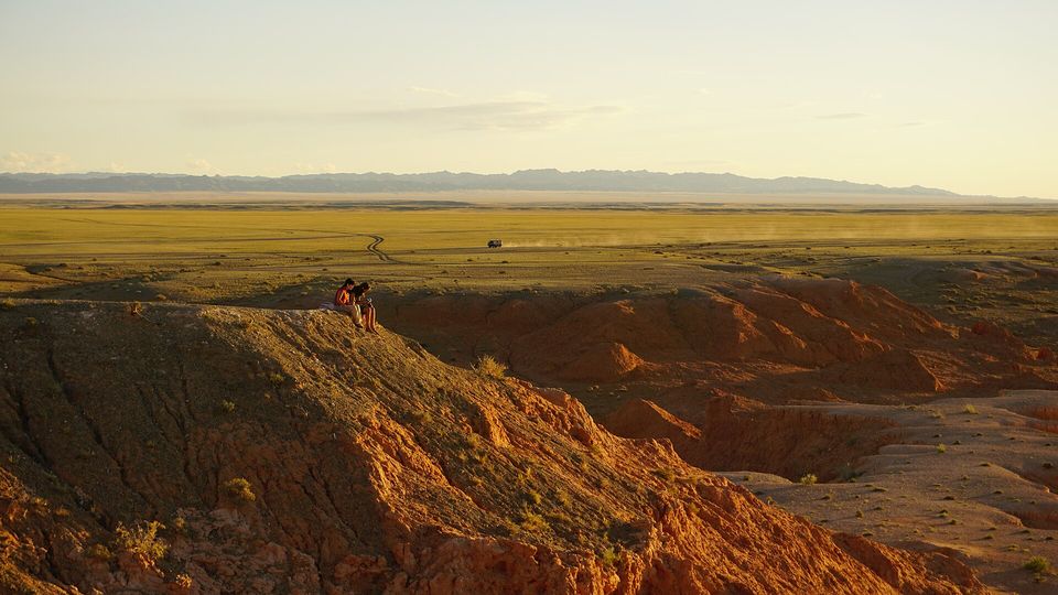 A grassy desert plain below red-orange cliffs, two small figures sitting on the ridge and a vehicle far off