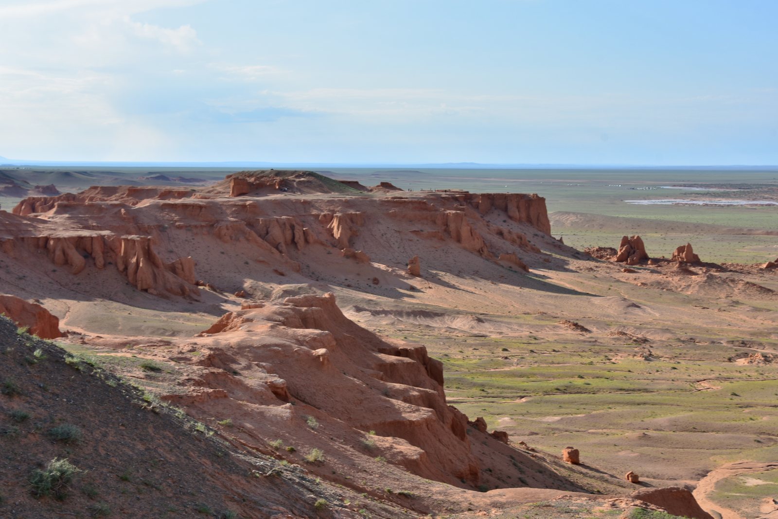 A band of eroded red-orange sandstone standing above the flat floor of the Gobi, gullied into buttresses, desert-steppe to the horizon