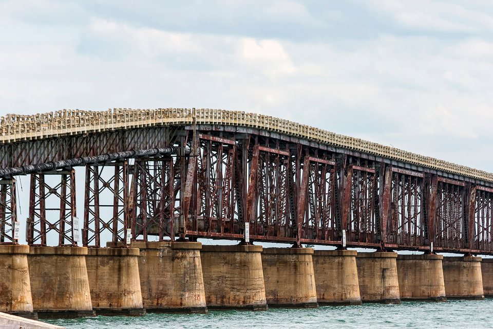 A rusted steel truss bridge on concrete piers over green water, a pale concrete road deck with railings laid along its top
