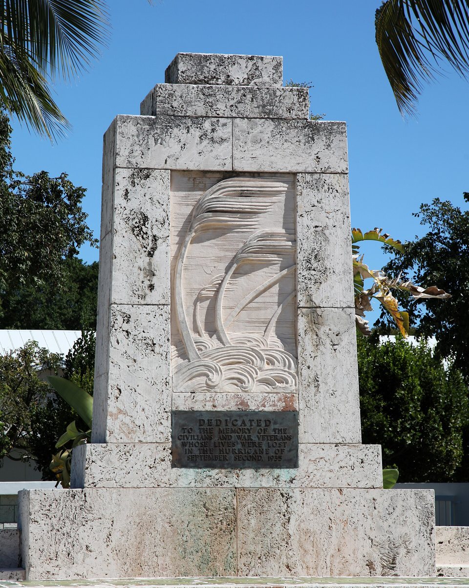A pale coral-stone monument carved with palms bending in wind above waves, a bronze dedication plaque below, blue sky behind