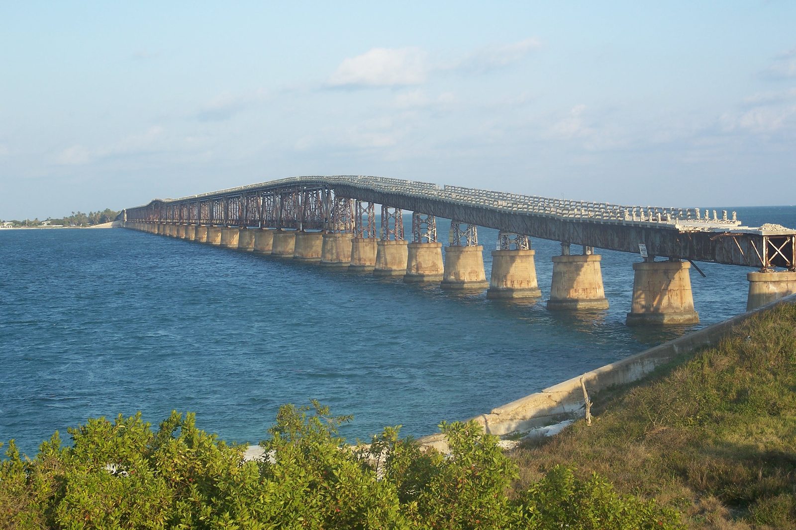 A rusted steel railway truss on concrete piers running out over turquoise water, a pale concrete road deck laid along its top, the deck breaking off at a removed span