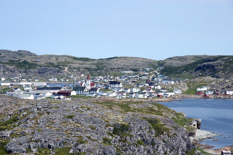 A harbour town of white and red wooden houses and a steepled church spreads across a rocky green valley beside a calm inlet, under a pale summer sky