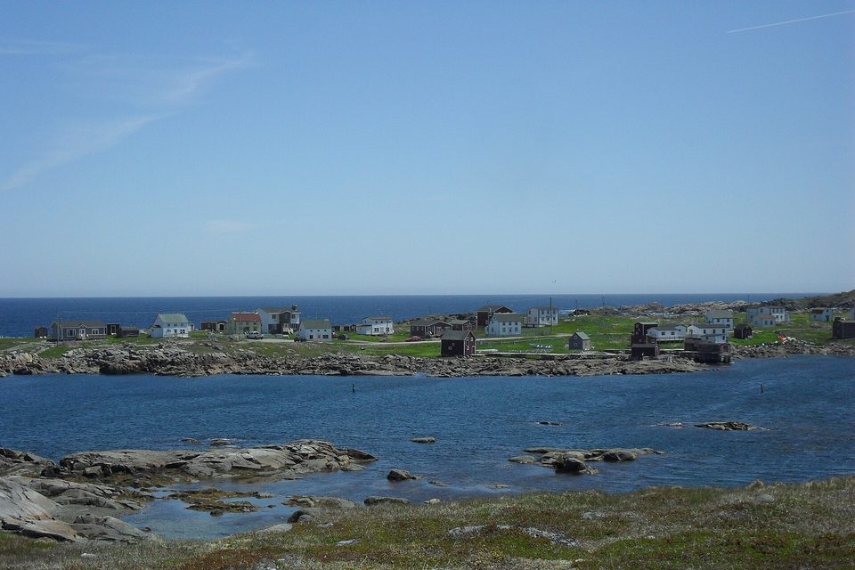 Small clapboard houses and dark fishing stages sit along a low shore across a rocky cove of deep-blue water, under a clear sky