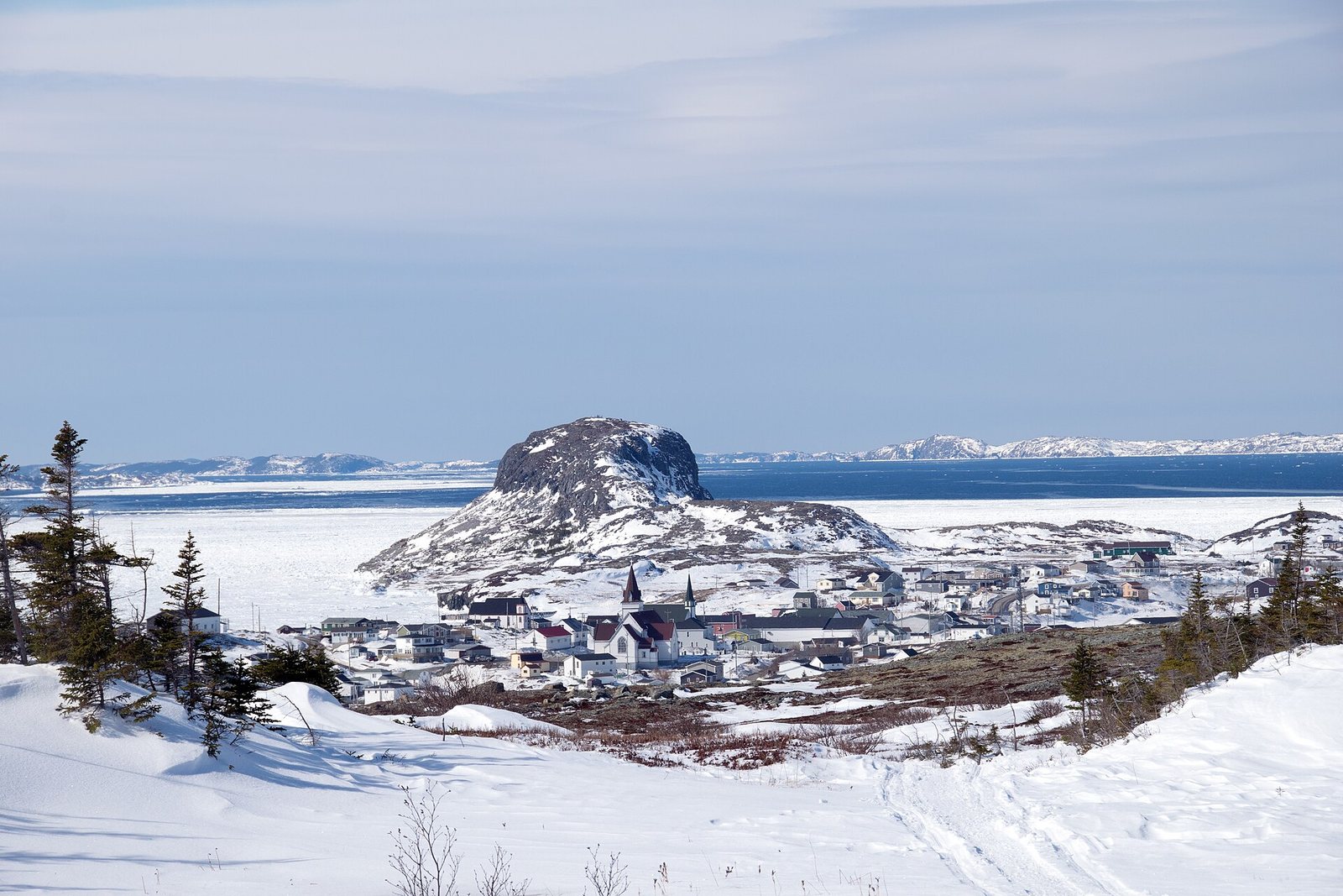 A dark dome of rock rises over a white clapboard town and its pointed church spire, pack ice on the bay and open blue water beyond, in clear light