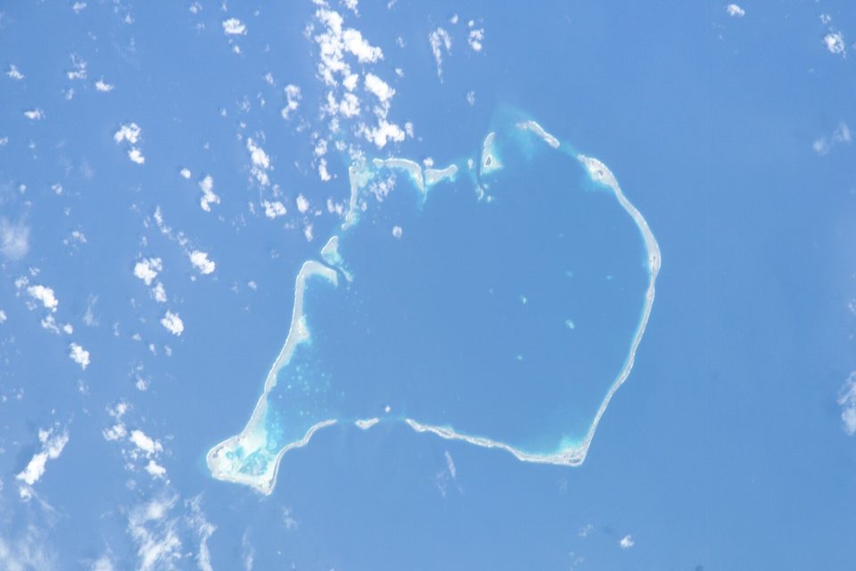 A photograph from orbit of a thin ring of reef and islets around a pale blue lagoon