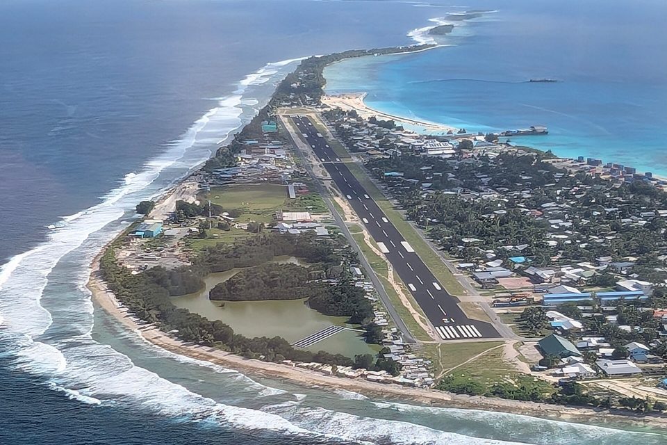 A runway running down the middle of a narrow atoll strip, with surf breaking on one side and a turquoise lagoon on the other, houses along its edges