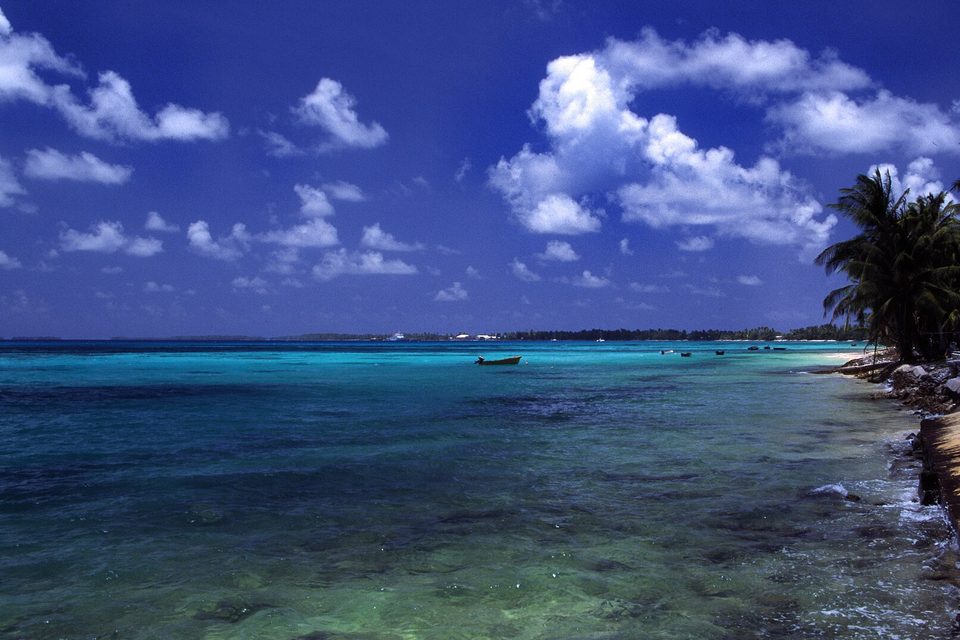 Clear shallow water over pale sand with moored boats and a seawall on the lagoon side