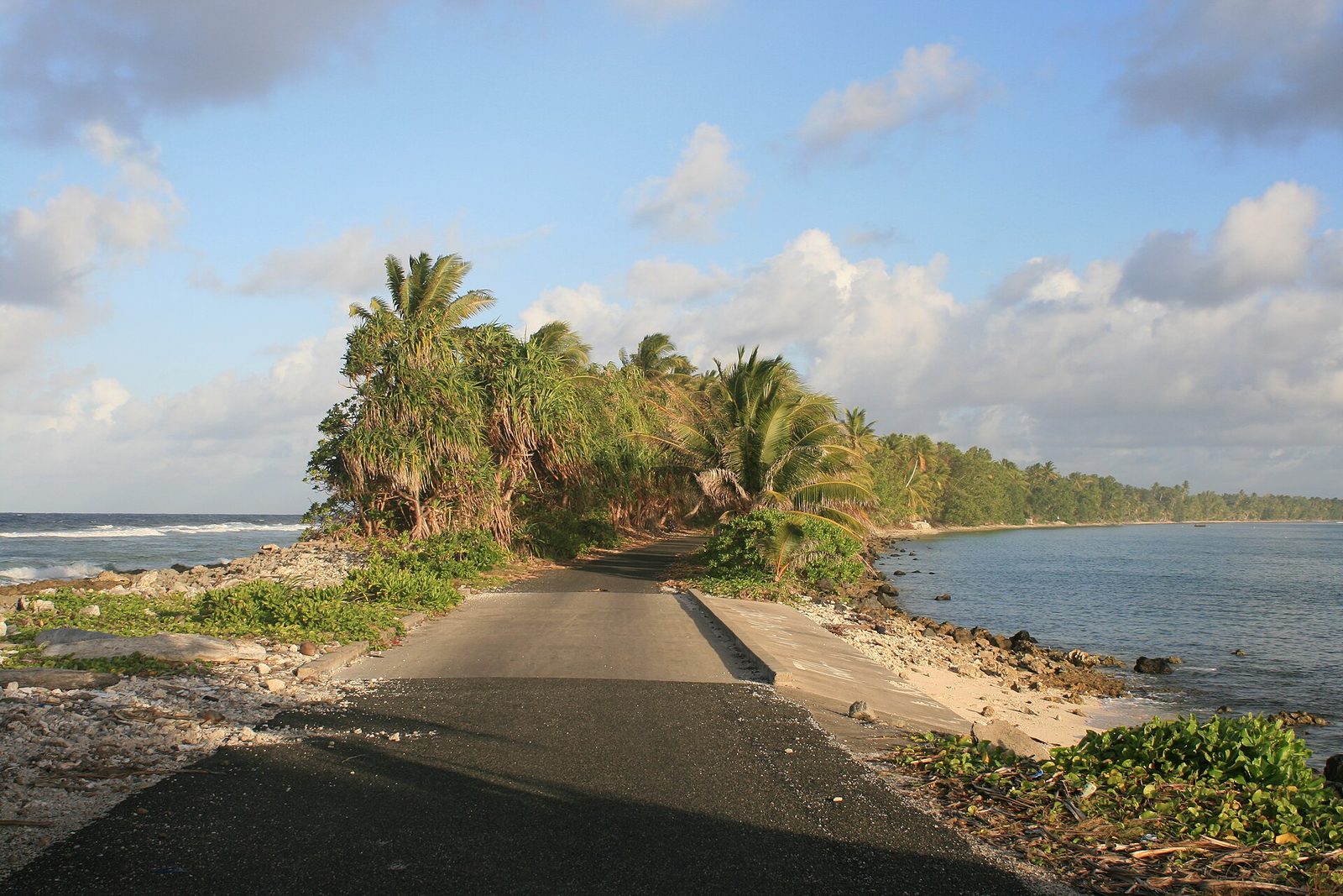 A single-lane road running between pandanus and palms with the ocean on the left and the lagoon on the right, in low gold light