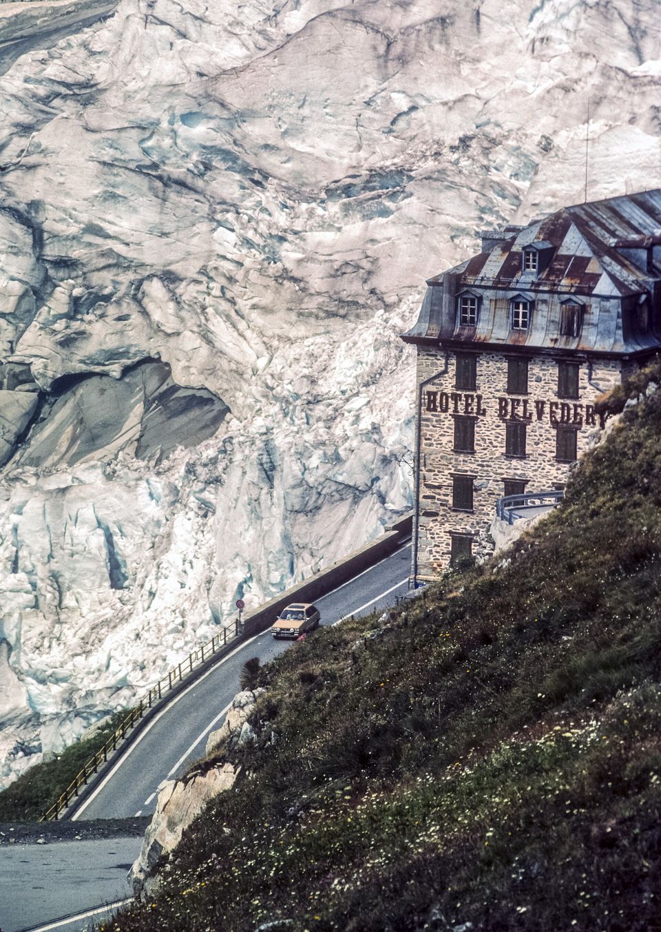 A tall stone hotel with a steep metal roof on a hairpin of the road, the cracked white ice of the Rhone Glacier filling the frame behind it, one small car on the bend