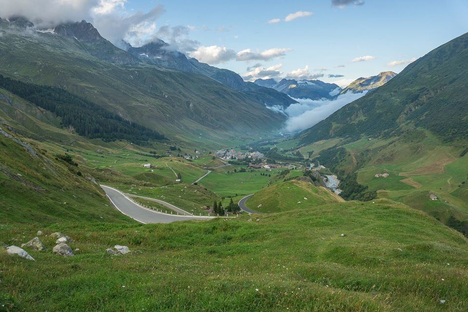 A road curving down a grassy mountainside toward a village on a green valley floor, cloud boiling out of a side valley, dark peaks behind