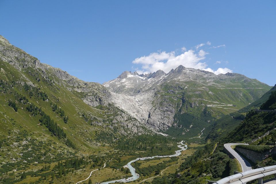A pale blue river braiding through a green valley floor toward a grey rock step below jagged snowy peaks, a road hairpin and guardrail at the right