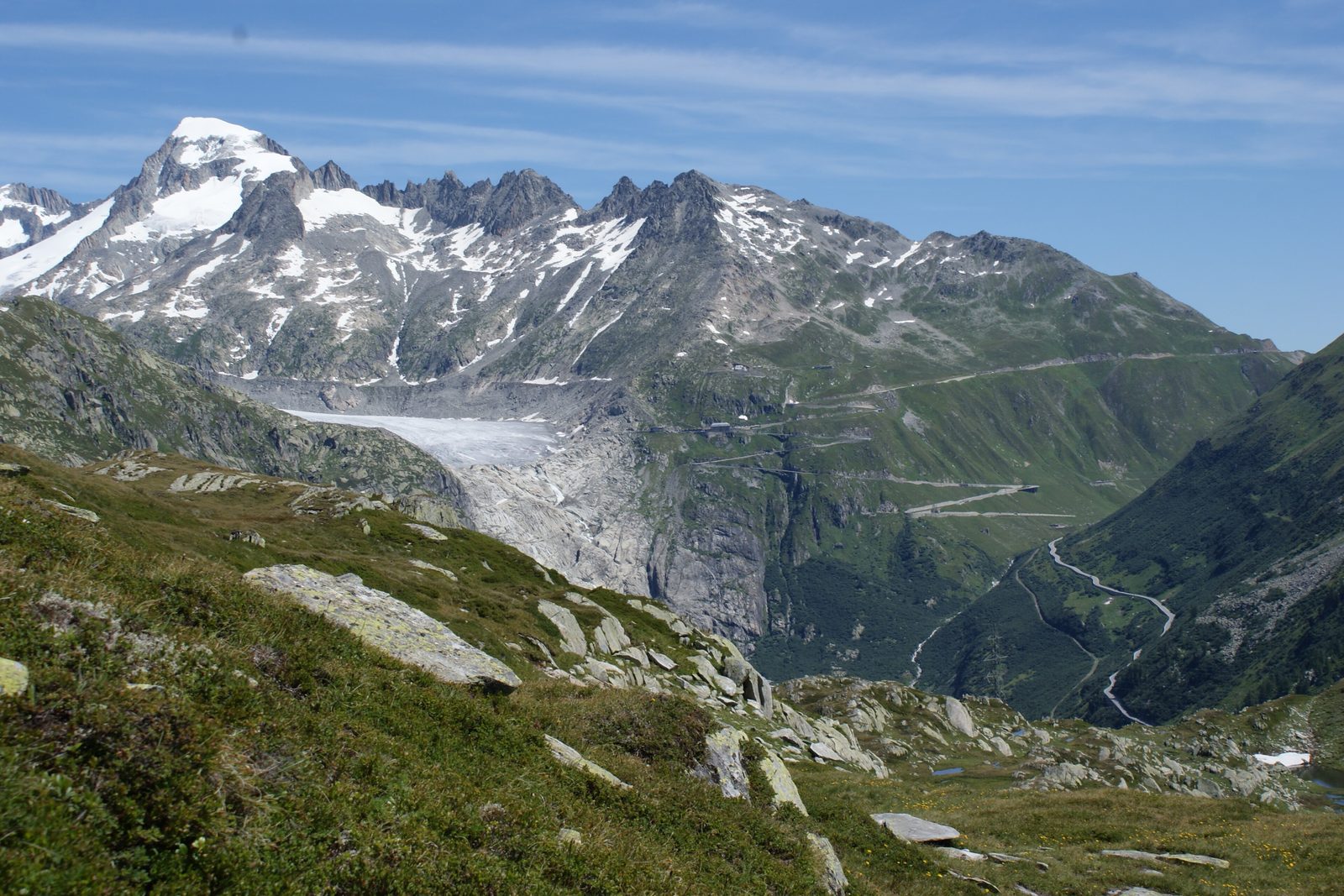 The Furka road zigzagging up a green mountainside beside the Rhone Glacier’s grey tongue, below the snowy Galenstock, seen across from the Grimsel side with alpine turf in the foreground