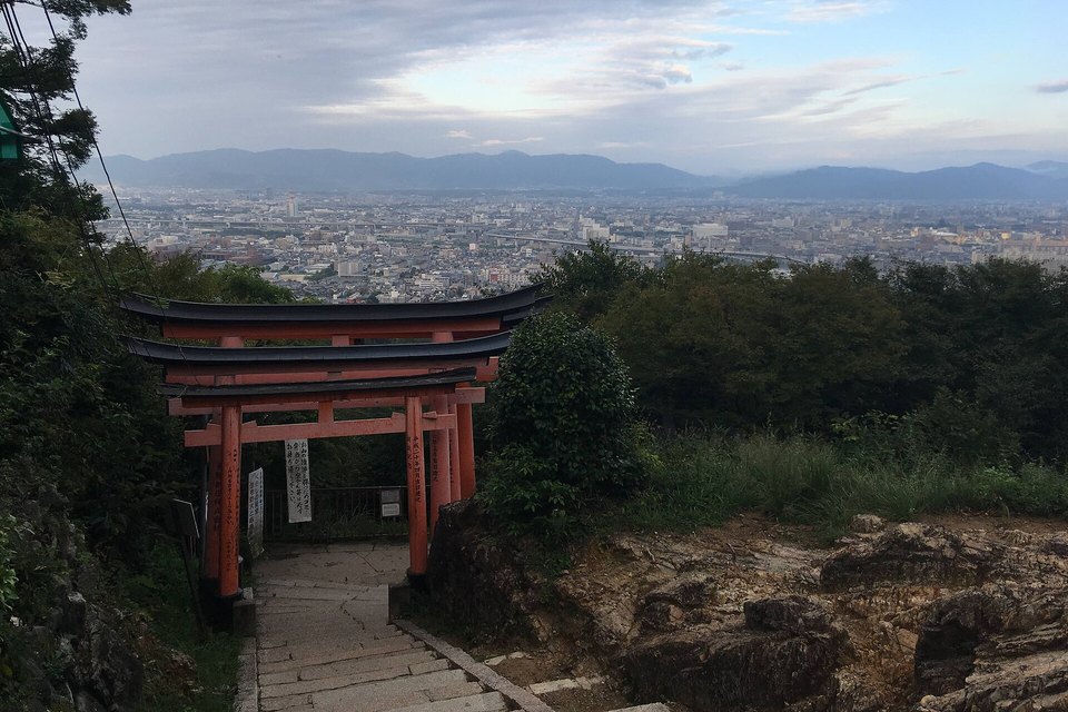 A stone stair descending through a red torii gate toward a hazy city plain and distant hills under a cloudy sky