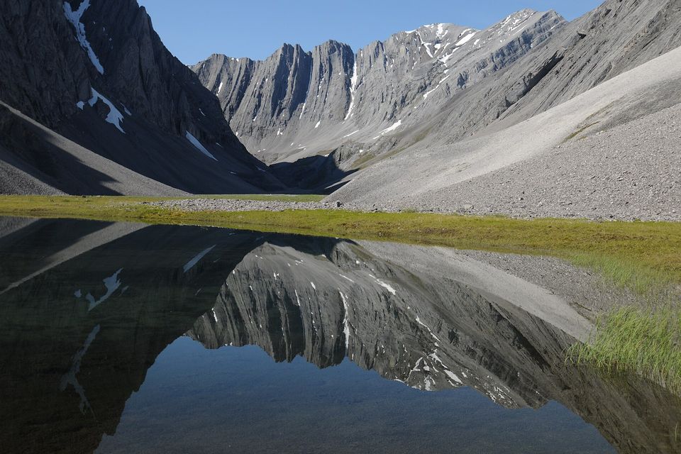 Grey limestone mountains reflected in a still pond below a green slope and scree