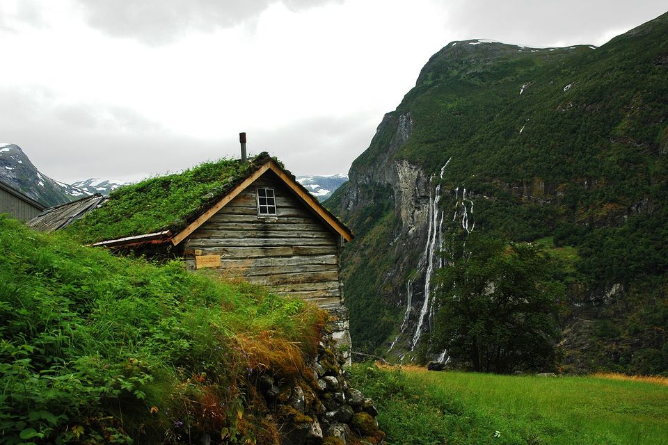 A log house with a turf roof on a green slope, a tall waterfall in several streams down a cliff beyond