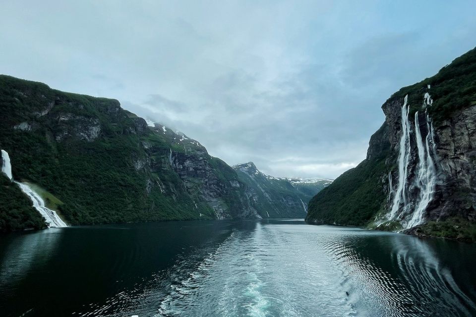 Two waterfalls on facing cliffs seen over a boat wake under a grey sky