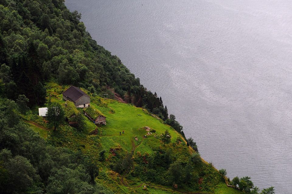 A green shelf with a few farm buildings high above a grey fjord, forest around it and two tiny walkers on the meadow
