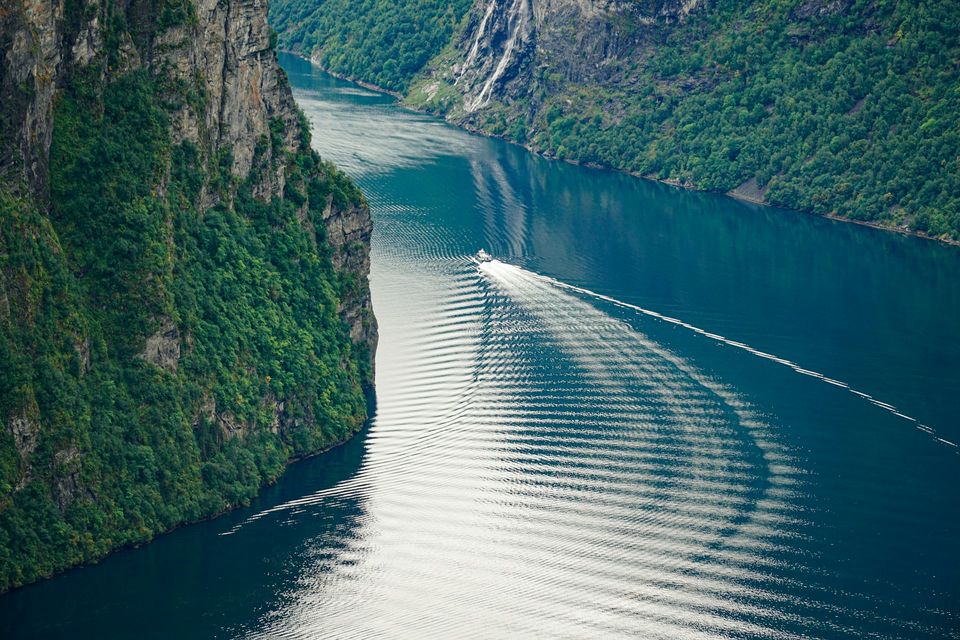 A boat wake fanning across dark fjord water below forested slopes, a waterfall at the top right