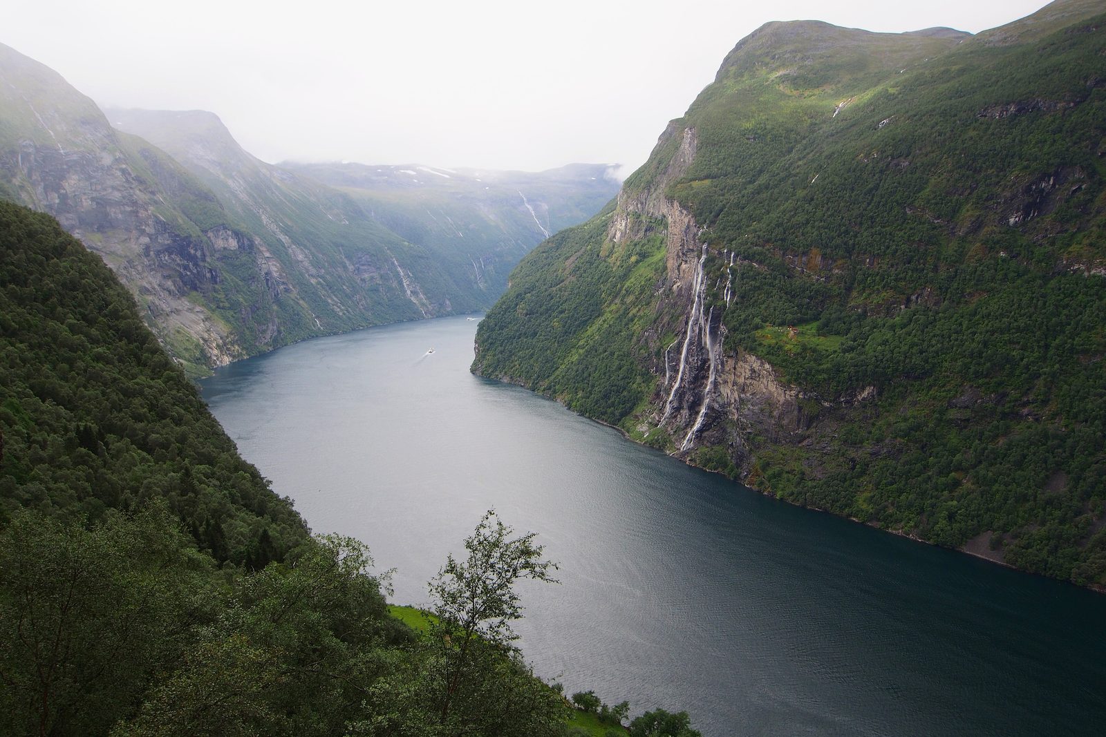 A dark fjord running between steep green walls under mist, a waterfall in seven white ribbons on the right