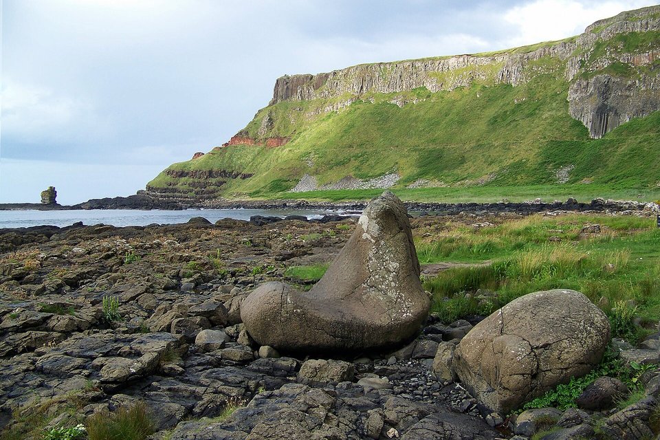 A large grey boulder shaped like a boot lying among dark rocks on the shore, green cliffs with pale bands of columns rising behind it