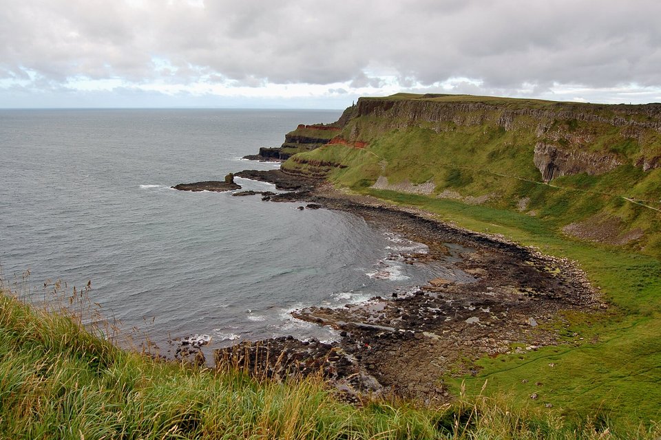 A curved bay of dark boulders under a grassy headland whose cliff face is made of vertical columns, a grey sea and a cloudy sky