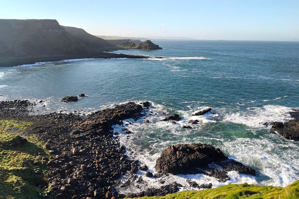 Surf breaking on dark boulders below a grassy clifftop, with headlands stretching away along the coast under a blue sky