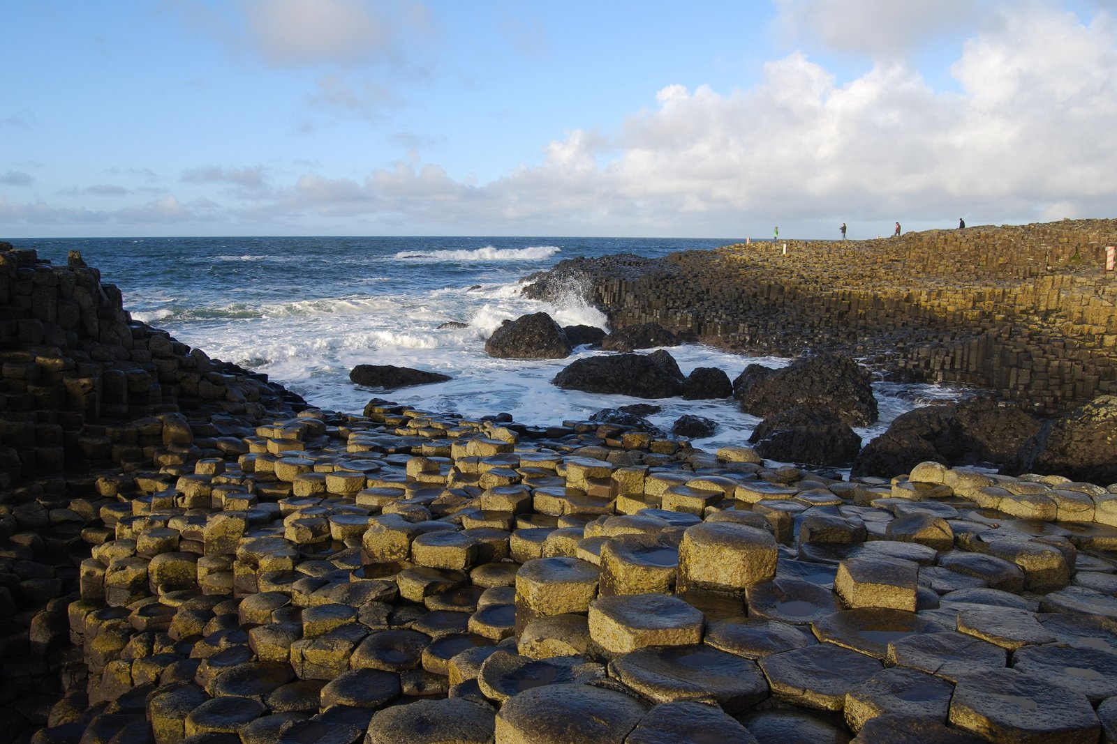 A pavement of hexagonal basalt columns with wet, gold-lit tops running down to a shore of breaking waves, taller columns forming a cliff on the right, under broken cloud