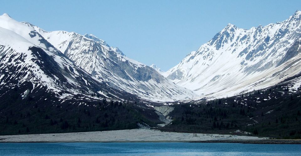 A wide U-shaped valley between snowy mountains opening onto a gravel shore and blue water