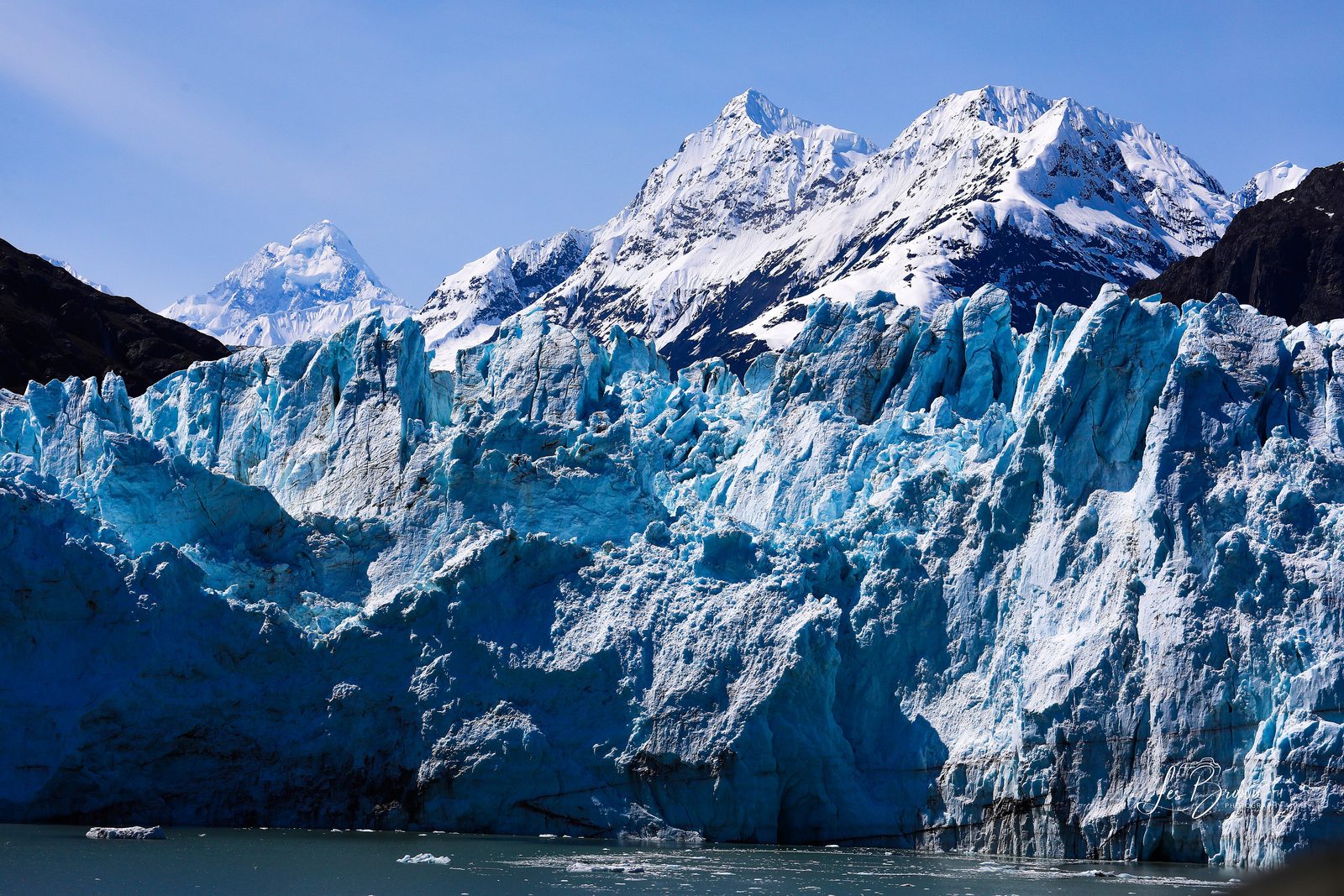 A wall of blue and white glacier ice meeting the water, with sharp snow-covered peaks above it under a blue sky