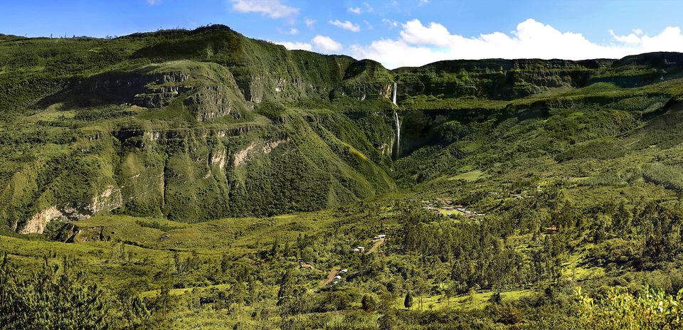 A wide green valley of pasture and scattered houses beneath a long cliff, with a thin waterfall falling in two drops at the head of the valley