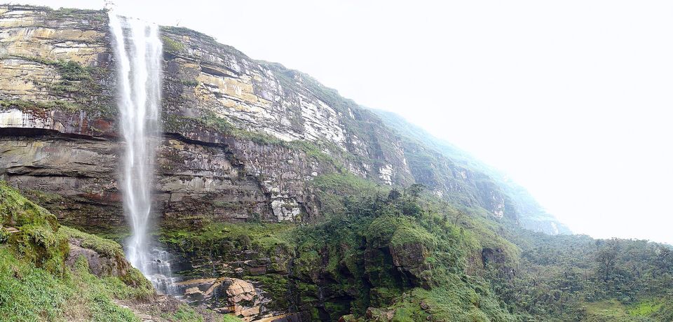 A tall thin waterfall falling down a wall of layered sandstone streaked with moss, with a pale hazy sky at the right