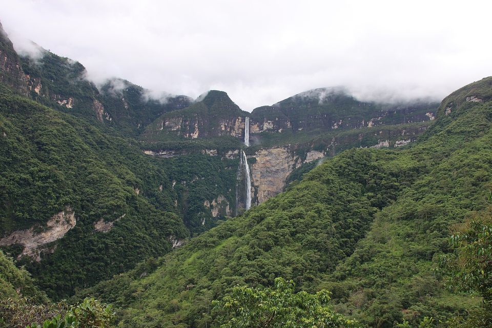A green forested gorge under low cloud that hides the cliff rim, with a thin waterfall falling in the middle distance