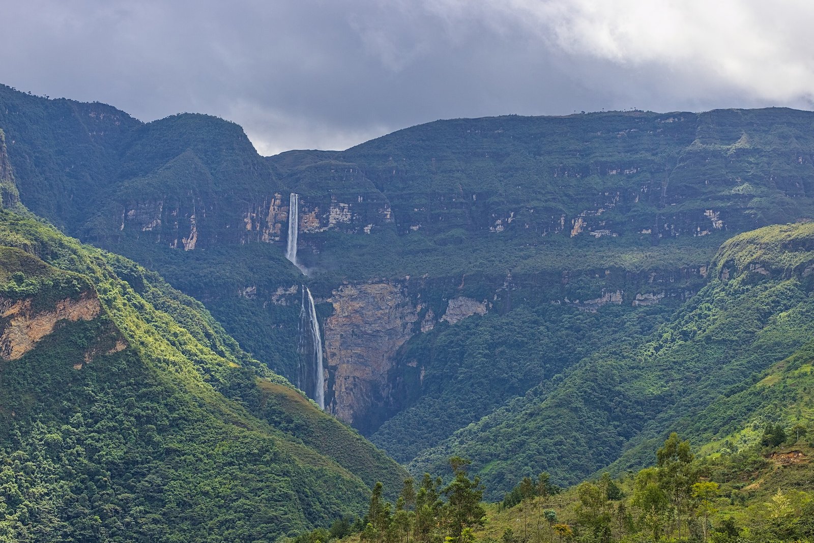 Two thin white waterfalls dropping one after the other from a layered sandstone cliff into a green forested gorge, under heavy grey cloud