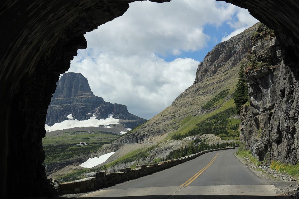 The view out of a dark rock tunnel onto a two-lane road edged with a low stone wall, curving along a steep mountainside toward a flat-topped peak with snowfields