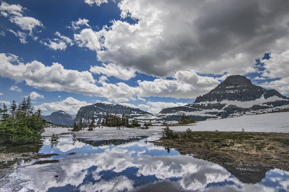 A shallow meltwater pool reflecting heavy white clouds, snowfields and dark conifers around it, a pyramid-shaped mountain at the right