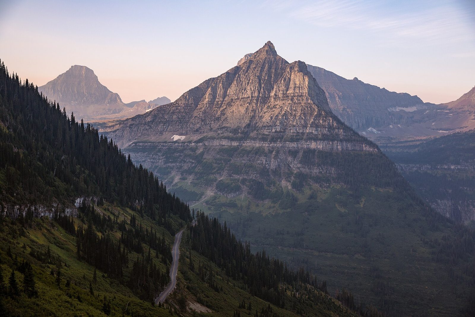 A pointed grey peak lit pink at sunrise above a deep forested valley, a narrow road cutting along the wooded slope below it and more peaks fading into haze behind