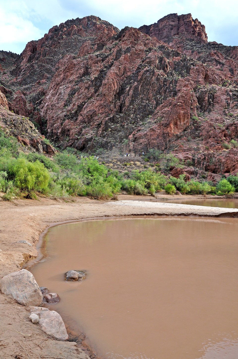 A brown river curving past a sandy beach and green shrubs below steep dark-red rock