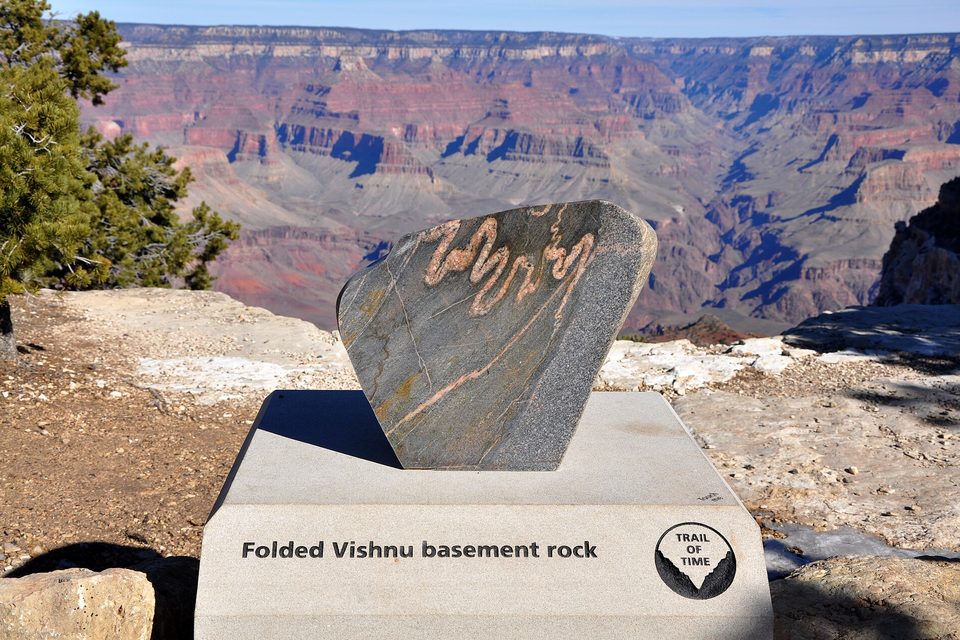 A tilted slab of dark banded rock mounted on a pale pedestal at a canyon rim, the canyon stretching behind it