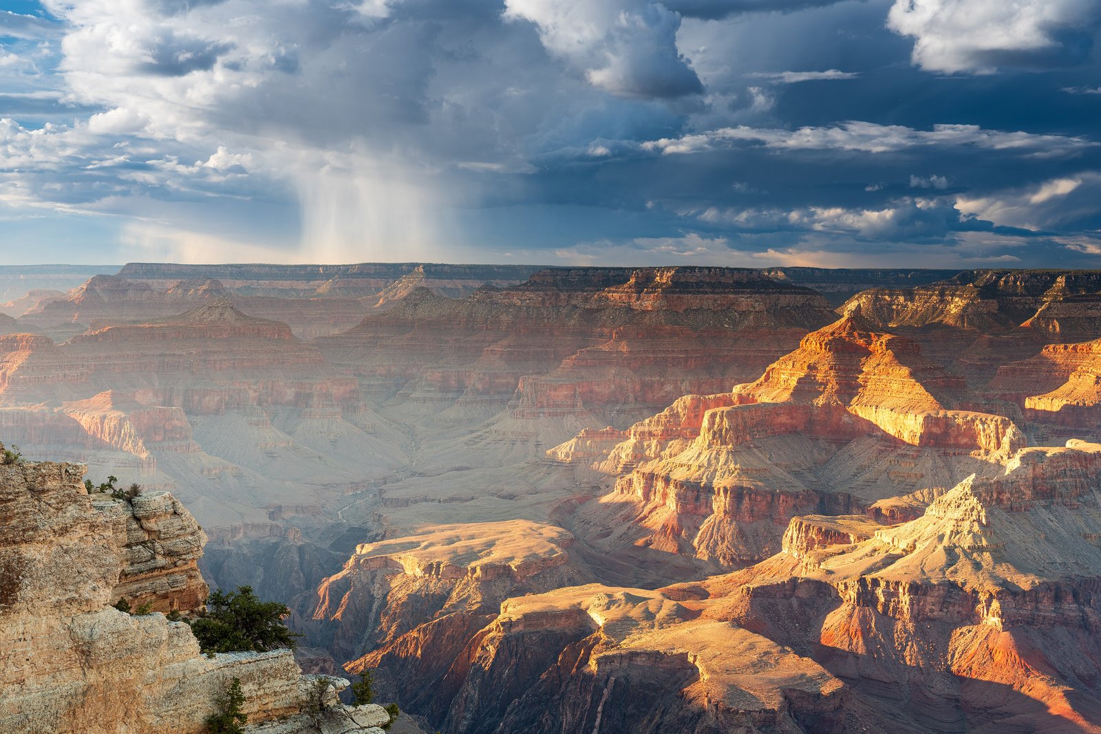A vast canyon of layered red and gold buttes lit by low sun beneath a dark cloud trailing rain, with a rock ledge at the left
