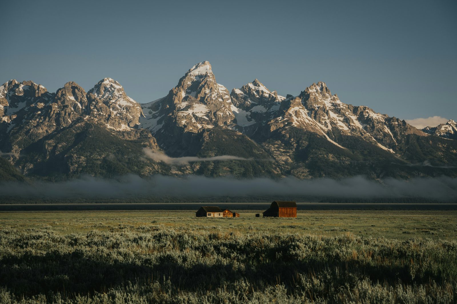 A jagged mountain range in first light above a bank of valley fog and a sage flat with two small barns