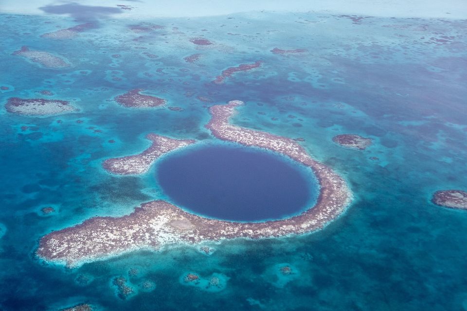 A wide aerial view of the round dark hole inside its ring reef, patch reefs scattered across the turquoise shallows around it