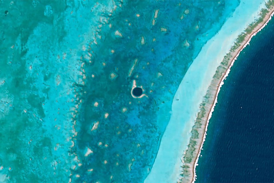 A satellite image of a shallow turquoise lagoon inside a reef, with a small dark round spot in the lagoon and open ocean beyond the reef crest
